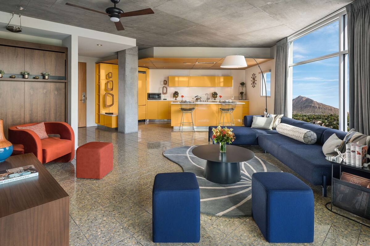 A modern living room with a sofa, ottomans, and a coffee table. There is a kitchen area with yellow cabinets and a large window.