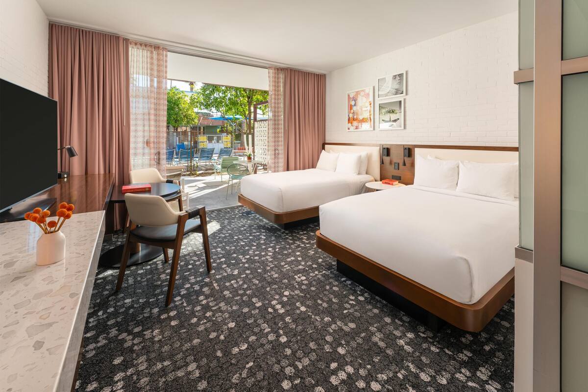 A hotel room with two beds, a desk with a chair, a TV, and a view of the outdoors.