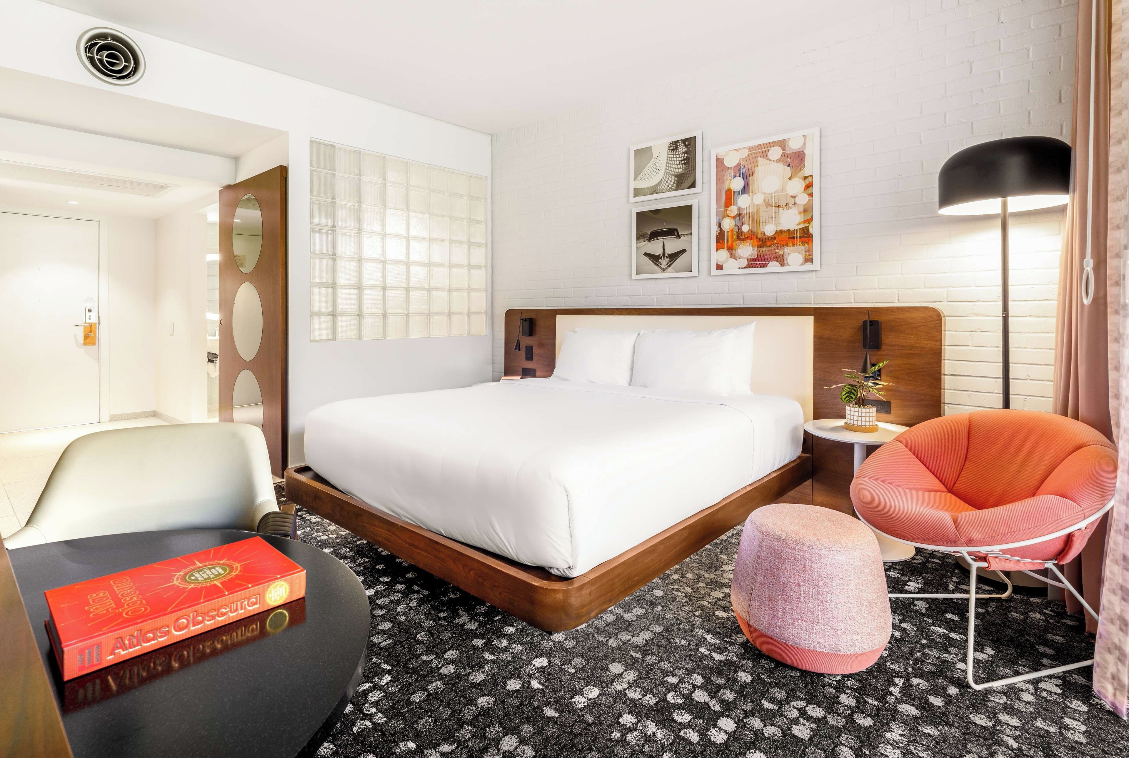 A hotel room with a bed, a chair, a small table, and a bookshelf.