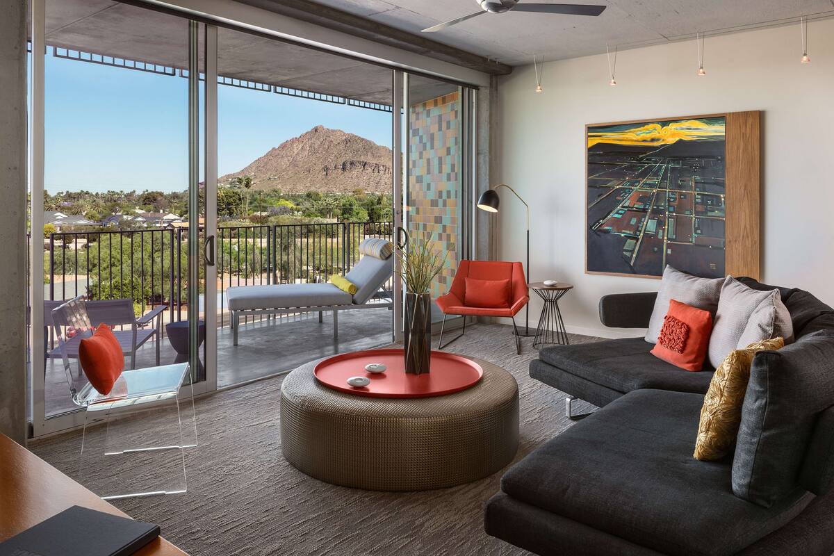 A modern living room with a large glass door leading to a balcony, a black sofa, a red ottoman, and a large abstract painting on the wall.