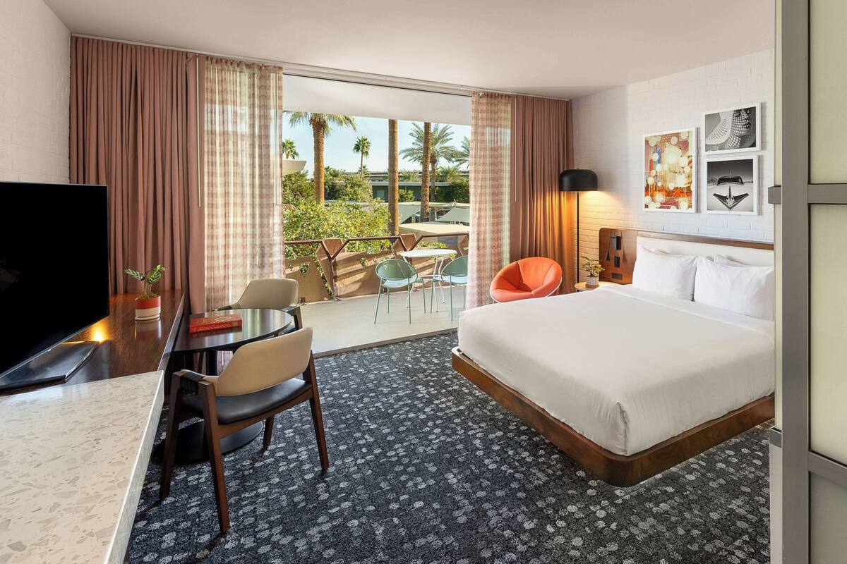 A hotel room with a bed, a desk with a TV, a chair, a table, and a balcony with a view of palm trees.