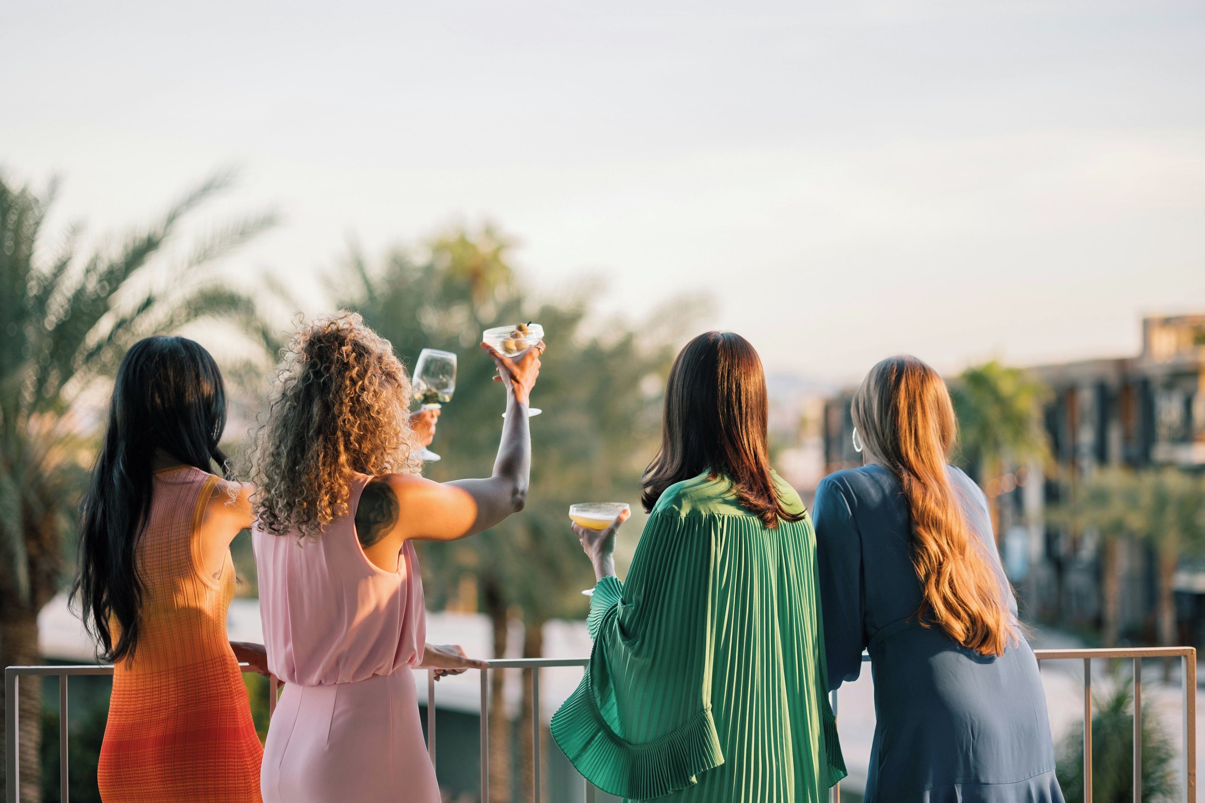 Un grupo de personas en un balcón con barandilla, sosteniendo bebidas, con vistas a un paisaje urbano con palmeras.