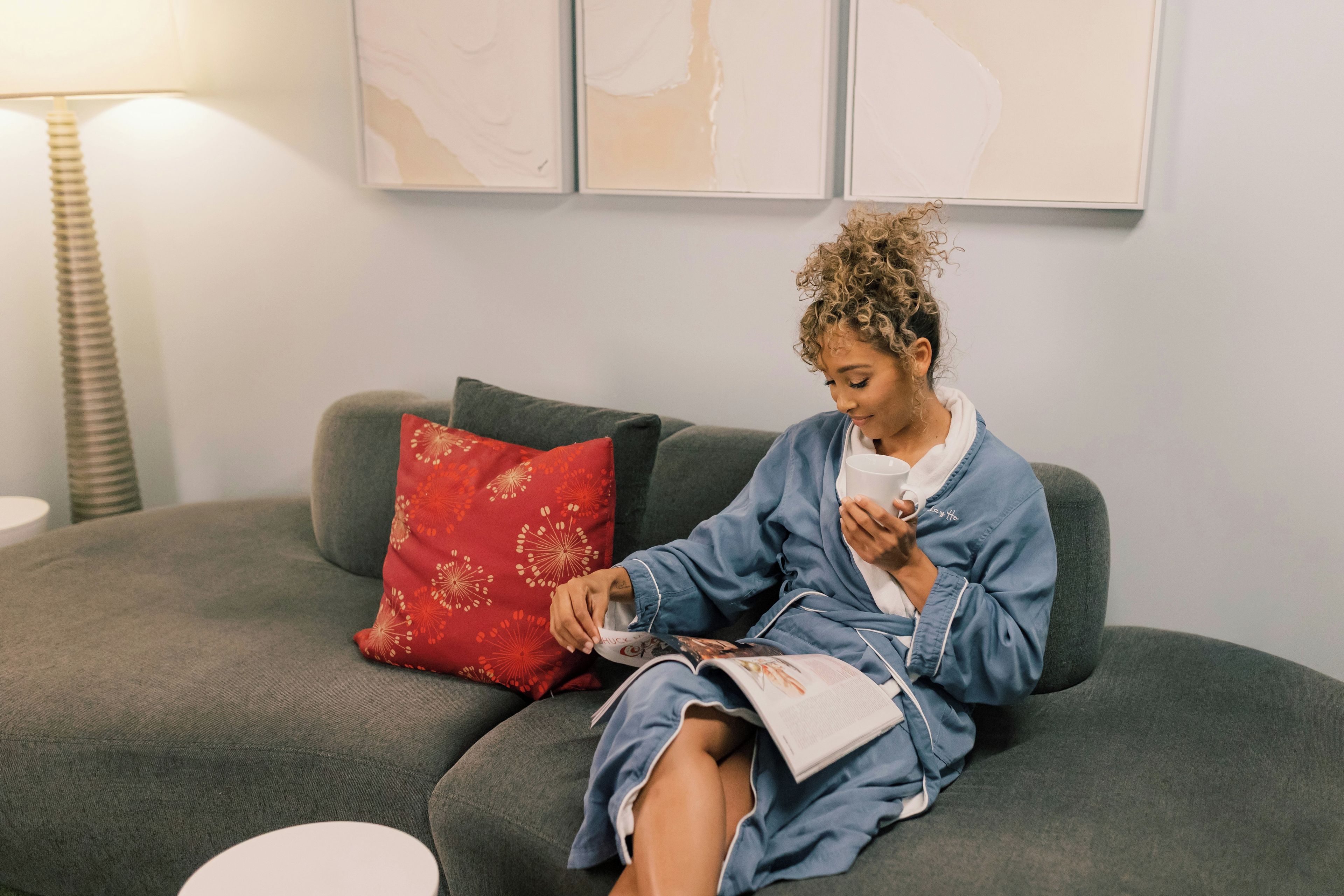 A person sitting on a grey sofa with a red pillow, holding a book and a cup.