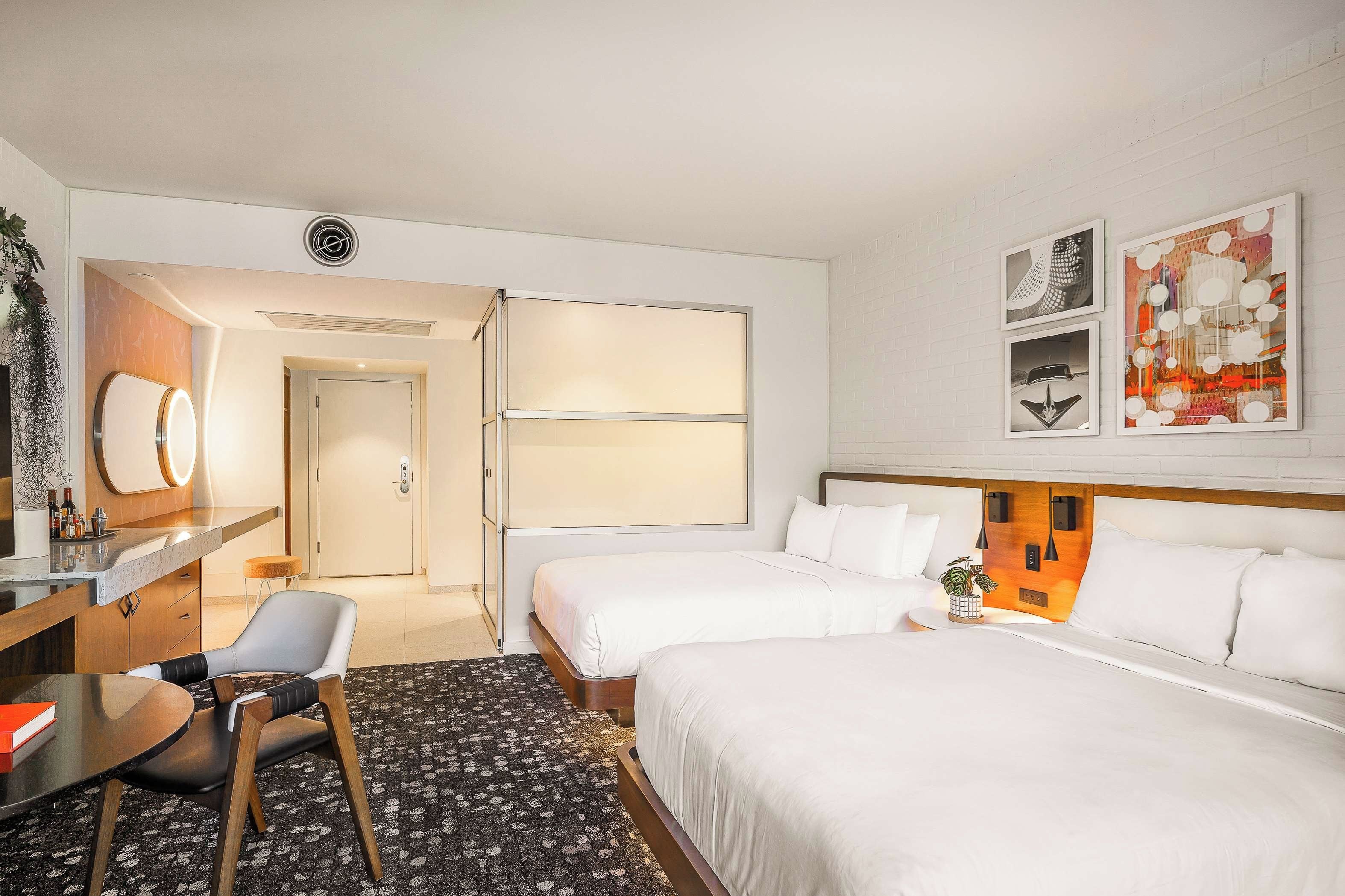 A hotel room with two beds, a desk with a chair, a bathroom with a shower, and artwork on the wall.