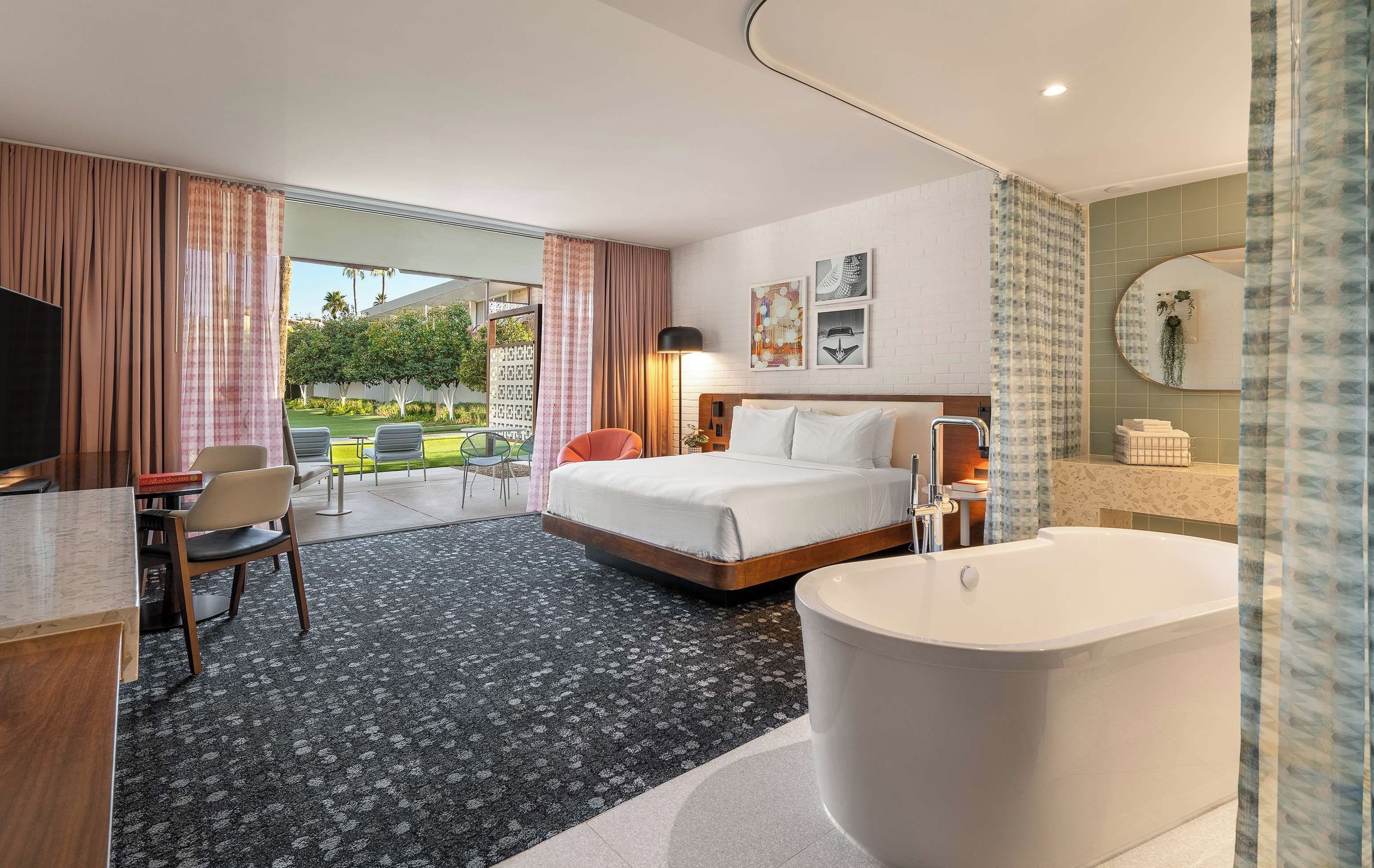 A hotel room with a bed, a desk, a chair, a bathroom with a bathtub, and a view of a garden.