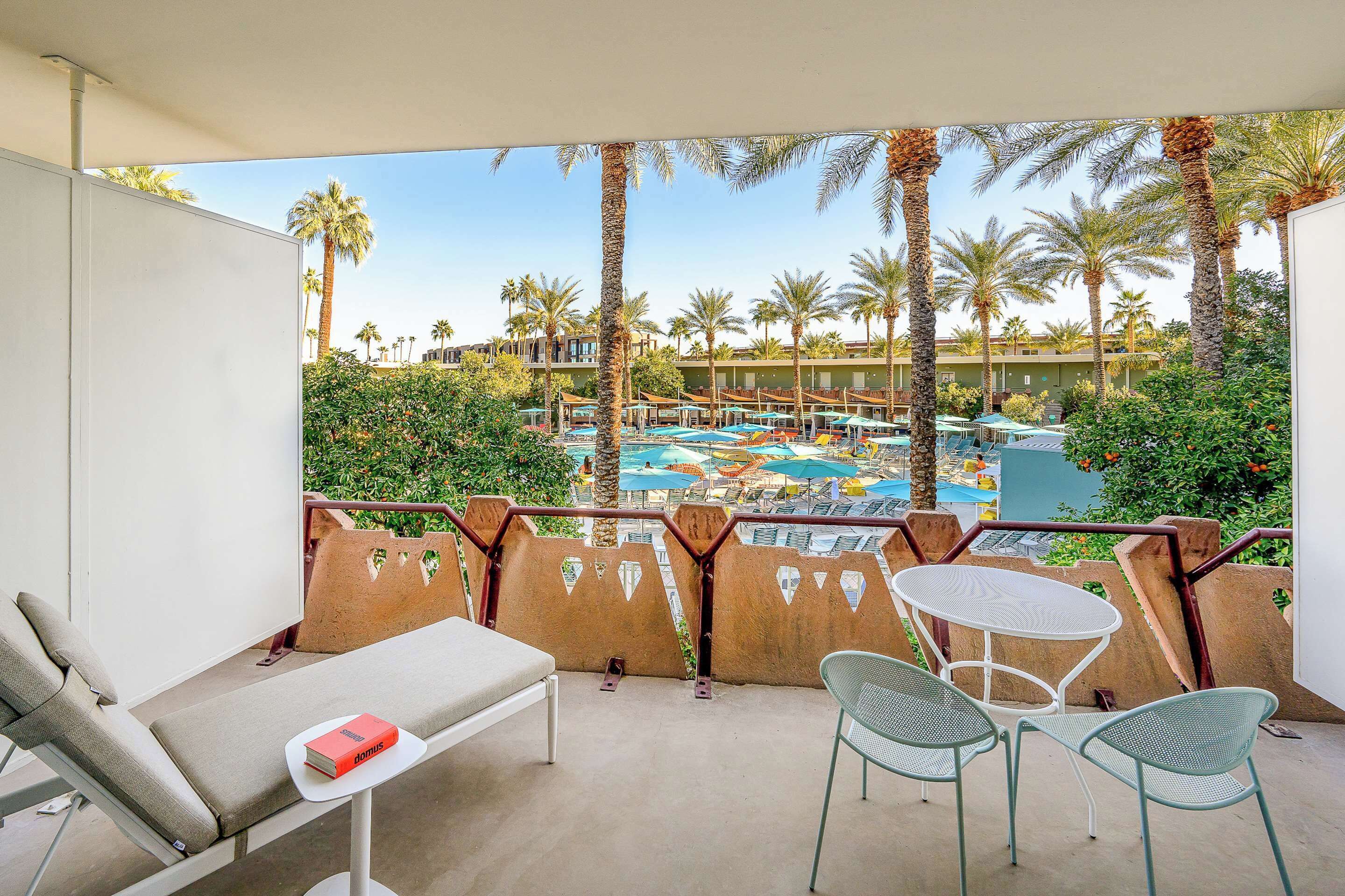 Un balcon équipé d’un fauteuil de relaxation, d’une petite table et de deux chaises. Il donne sur un espace piscine agrémenté de plusieurs parasols et de palmiers.