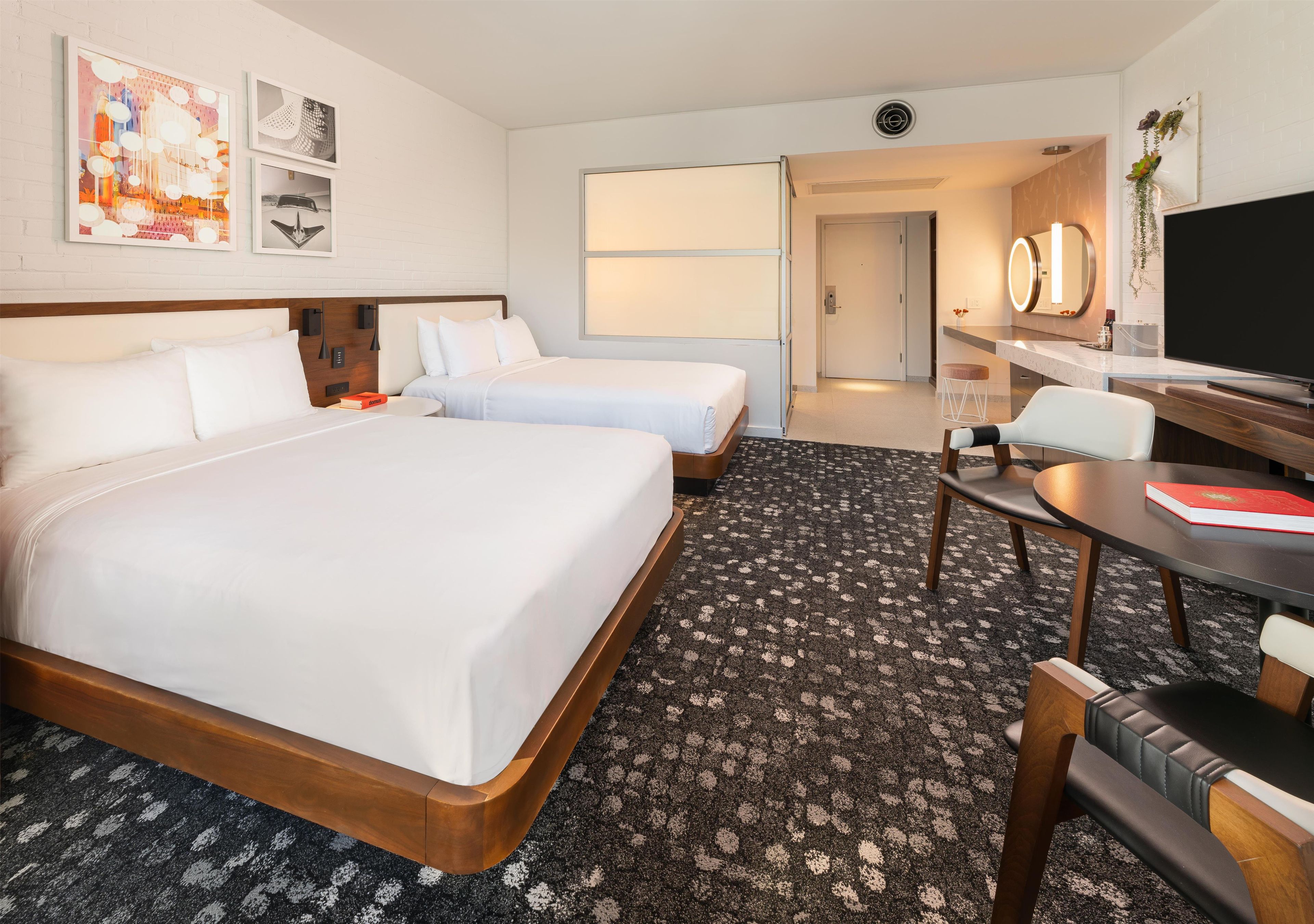 Habitación de hotel con dos camas, un escritorio, una silla, un televisor y un baño visible a través de una puerta abierta.