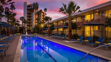 A pool area with lounge chairs and umbrellas, surrounded by palm trees and a building with balconies.