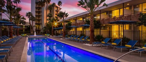 A pool area with lounge chairs and umbrellas, surrounded by palm trees and a building with balconies.