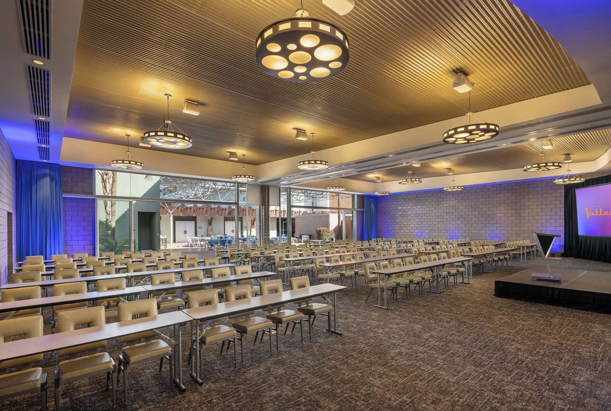 Une salle de conférence avec des rangées de tables et de chaises, une scène avec un écran et une grande fenêtre donnant sur l’extérieur.