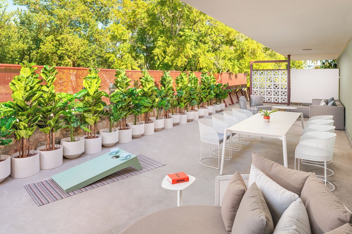 Un espace patio extérieur avec une table blanche, des chaises et un canapé. Des plantes en pot sont disposées le long du mur et un tapis à motifs recouvre le sol.