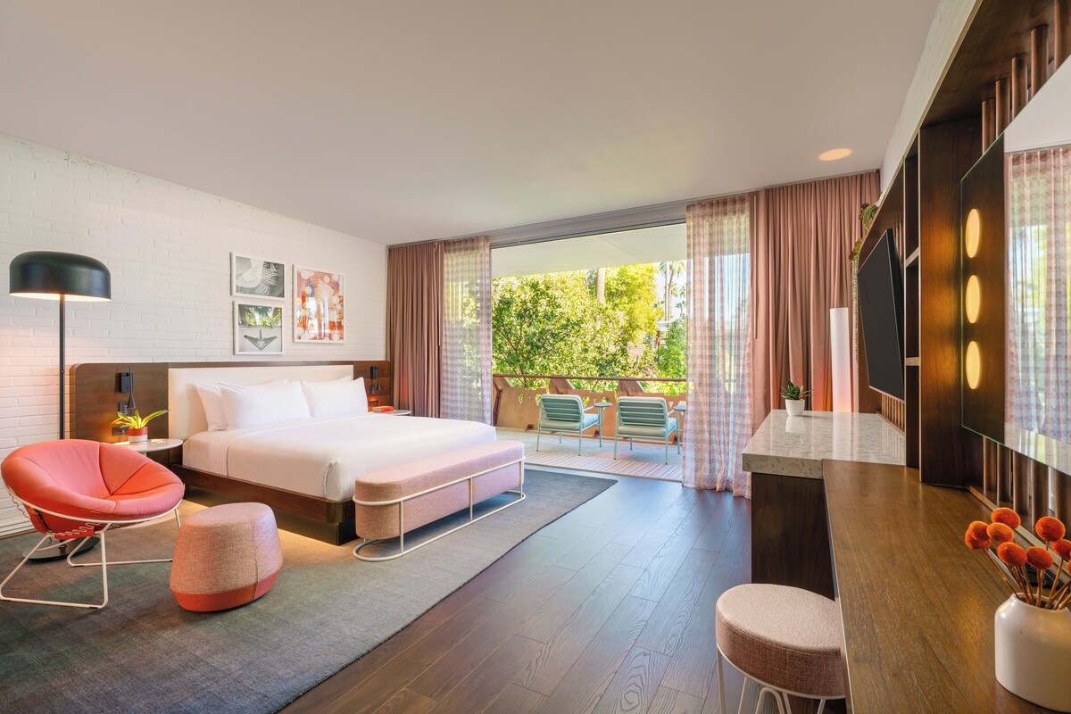 A modern hotel room with a large bed, a red chair, a small ottoman, a desk with a vase of flowers, and a balcony with a view of greenery.
