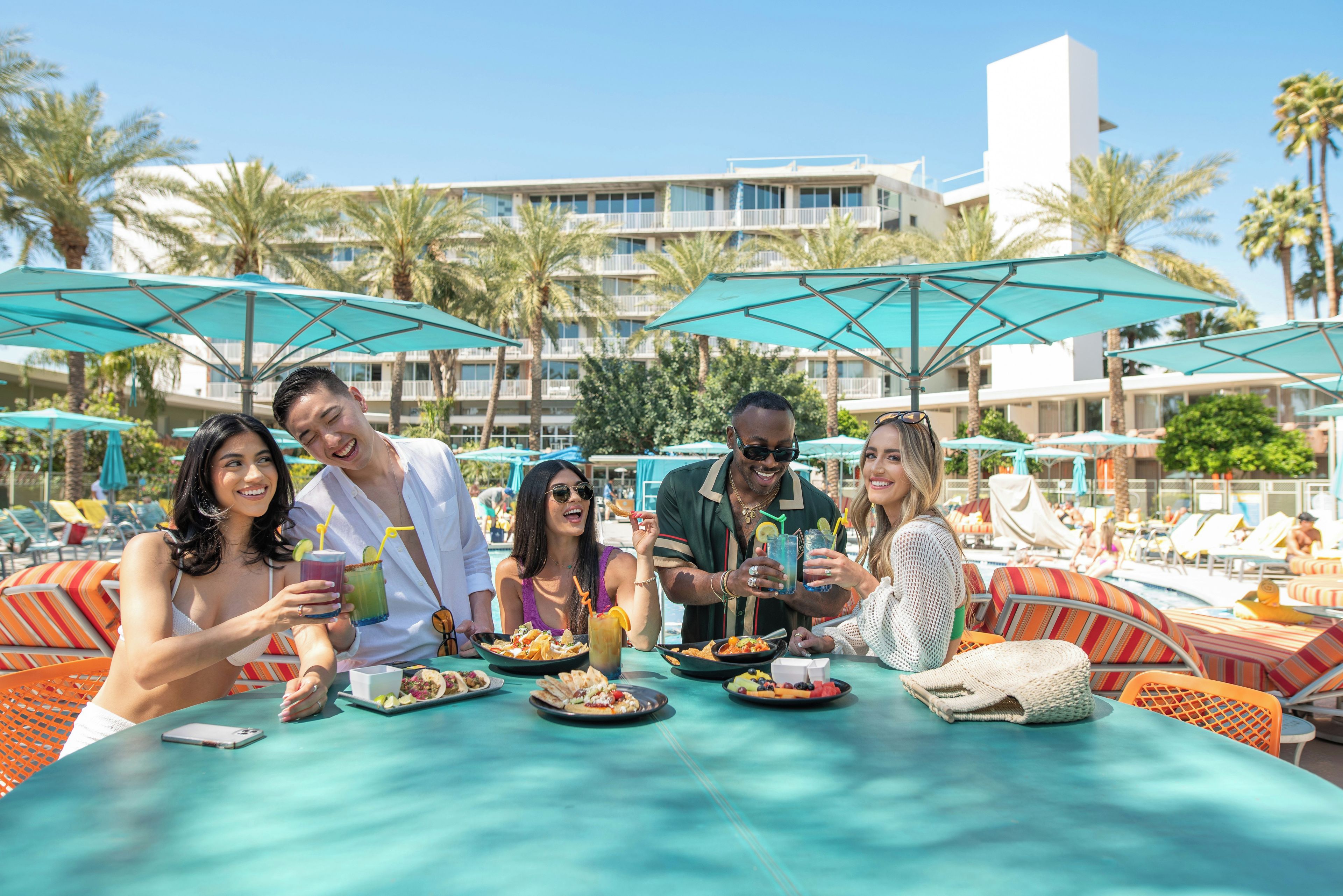 A group of people dining at an outdoor table with umbrellas, surrounded by palm trees and a hotel building in the background.