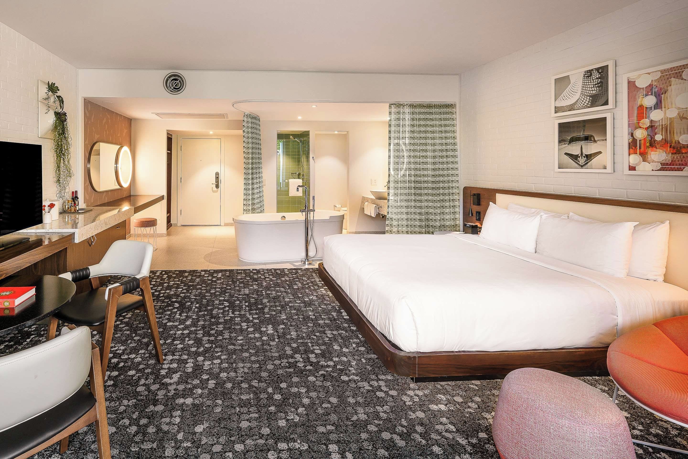 A hotel room with a large bed, a desk with a chair, a TV, a bathroom with a shower, and a small seating area.