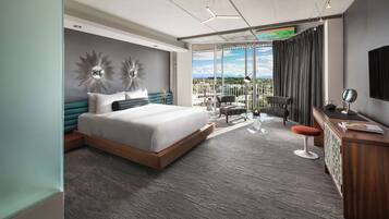 A modern hotel room with a large bed, a desk with a mirror, and a balcony with a view.