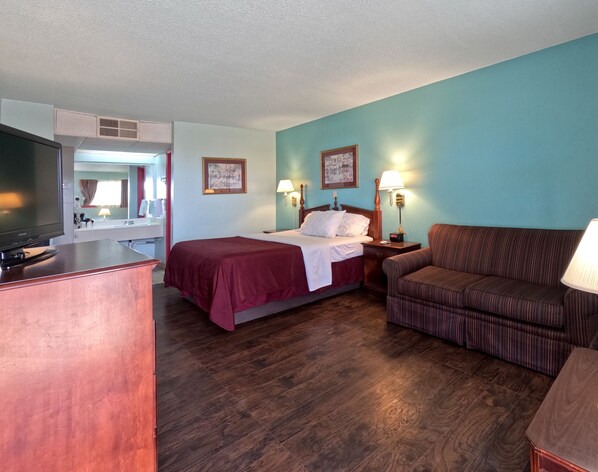 A hotel room with a bed, a sofa, a desk with a television, and a bathroom visible through a door.
