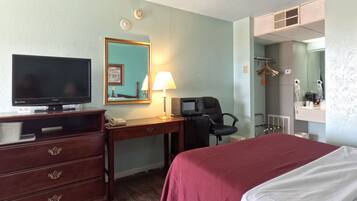 A hotel room with a bed, a desk with a computer, a television, a mirror, and a bathroom with a shower and toilet.