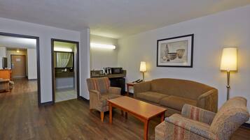 Habitación de hotel con sala de estar que incluye un sofá, dos sillones, una mesa de centro de madera y una mesa de comedor. Cuenta con una zona de cocina con refrigerador, microondas y una pequeña isla. Un cuadro enmarcado adorna la pared, y dos lámparas de pie se encuentran a cada lado del sofá.