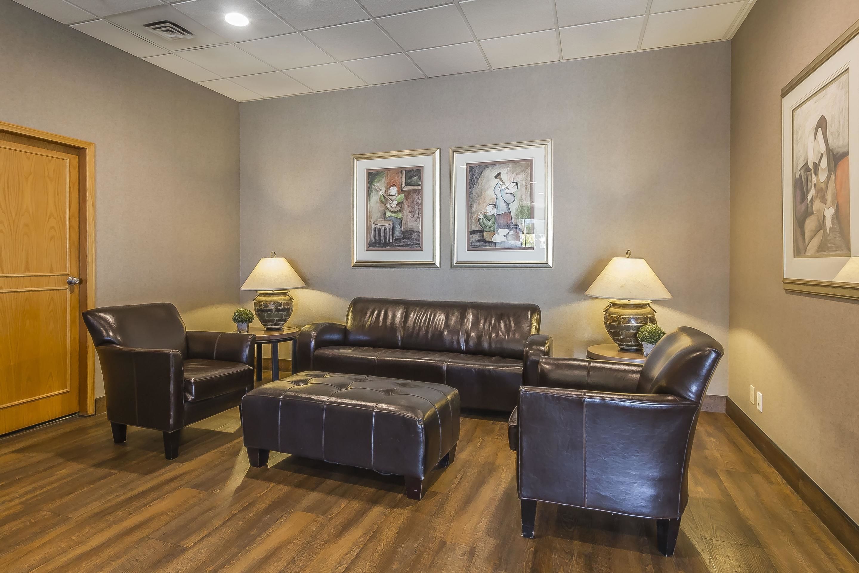 A lobby with a leather sofa, two armchairs, a matching ottoman, and two lamps on side tables. There are framed artworks on the wall and a wooden door on the left.