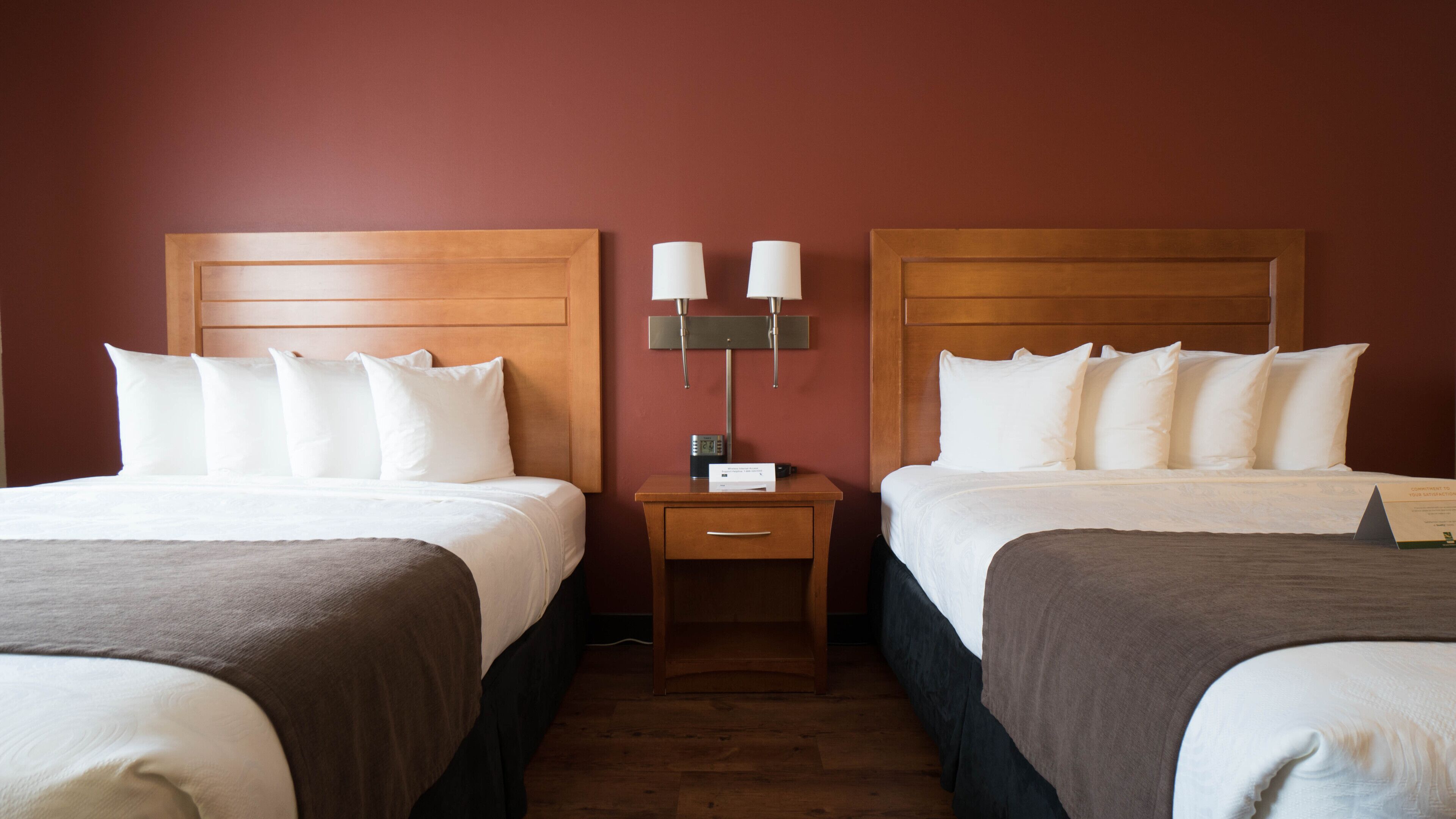 A hotel room with two beds, each with white linens and brown bedspreads. There is a wooden nightstand between the beds with a lamp and a clock. The walls are painted a warm red color.