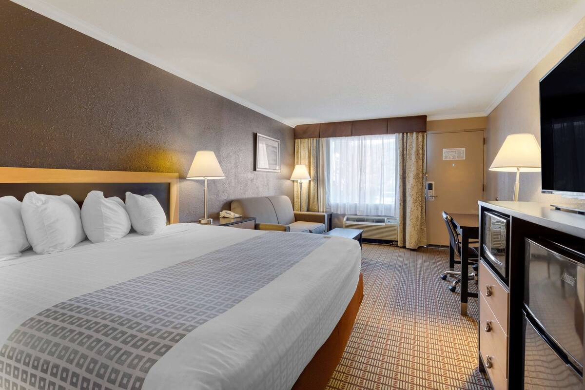 A hotel room with a large bed, two bedside tables with lamps, a desk with a chair, a television, and a window with curtains.