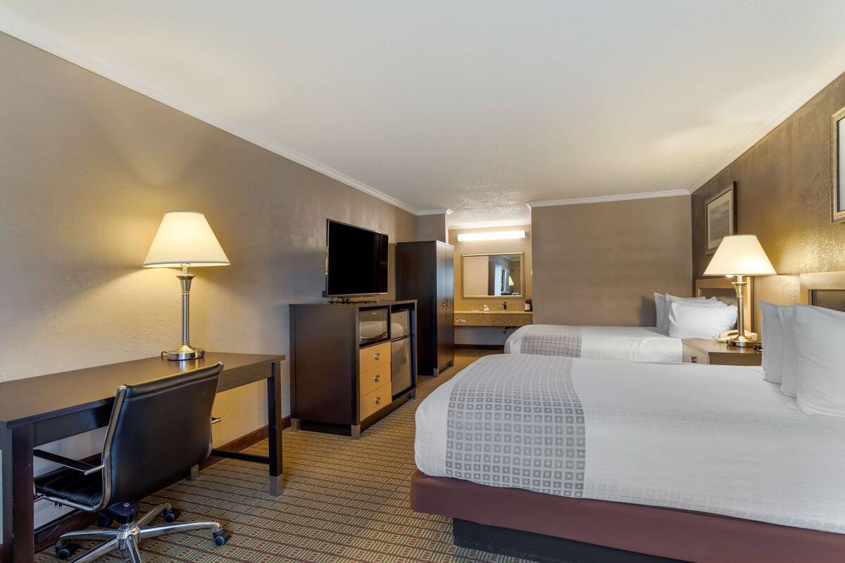 A hotel room with a desk, two beds, a TV, and a bathroom with a mirror.
