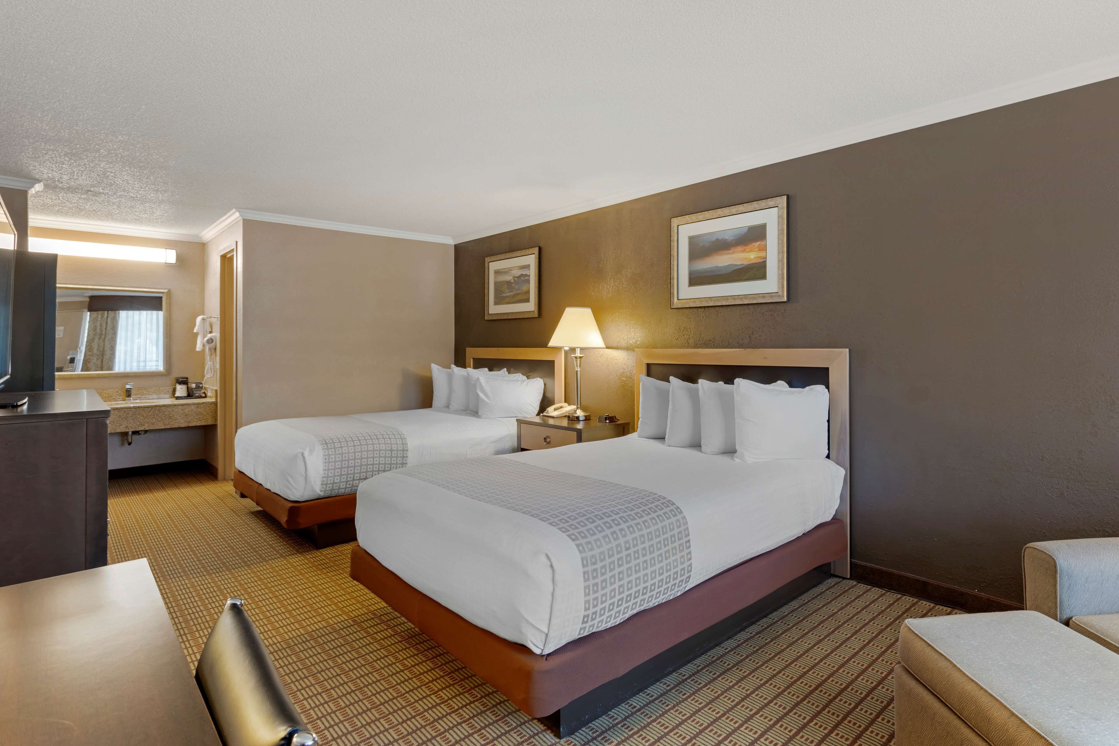 A hotel room with two beds, a desk, a chair, a lamp, and a painting on the wall.