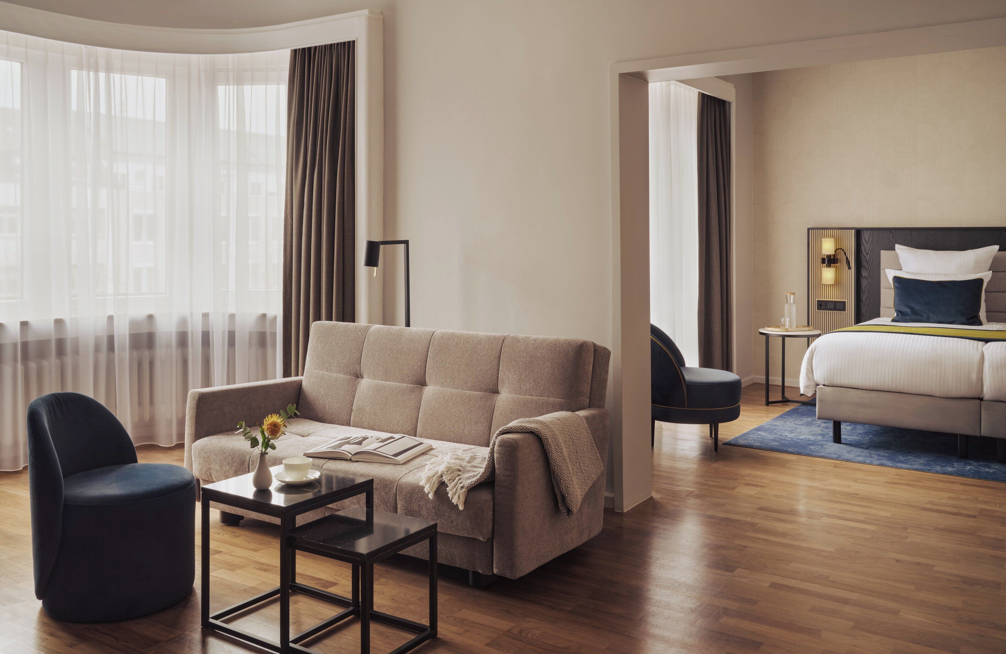 A modern hotel room with a sofa, armchair, and a bed with a blue accent pillow.