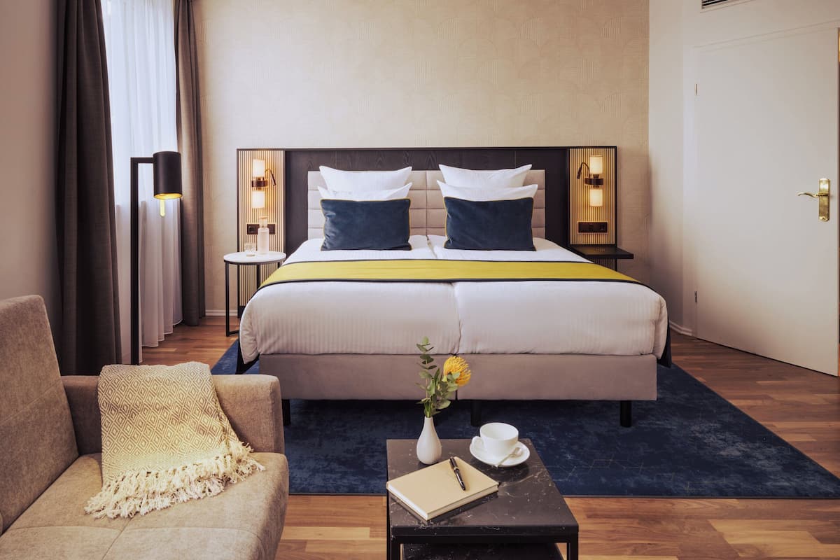 Quarto de hotel com uma cama grande, duas mesas de cabeceira com luminárias, um sofá, uma mesa de centro com um livro e um vaso de flores.