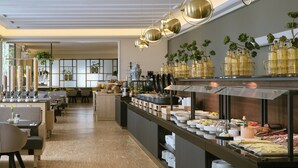 Un restaurante moderno con una larga barra que exhibe diferentes alimentos, una zona de estar con mesas y sillas, y plantas decorativas.