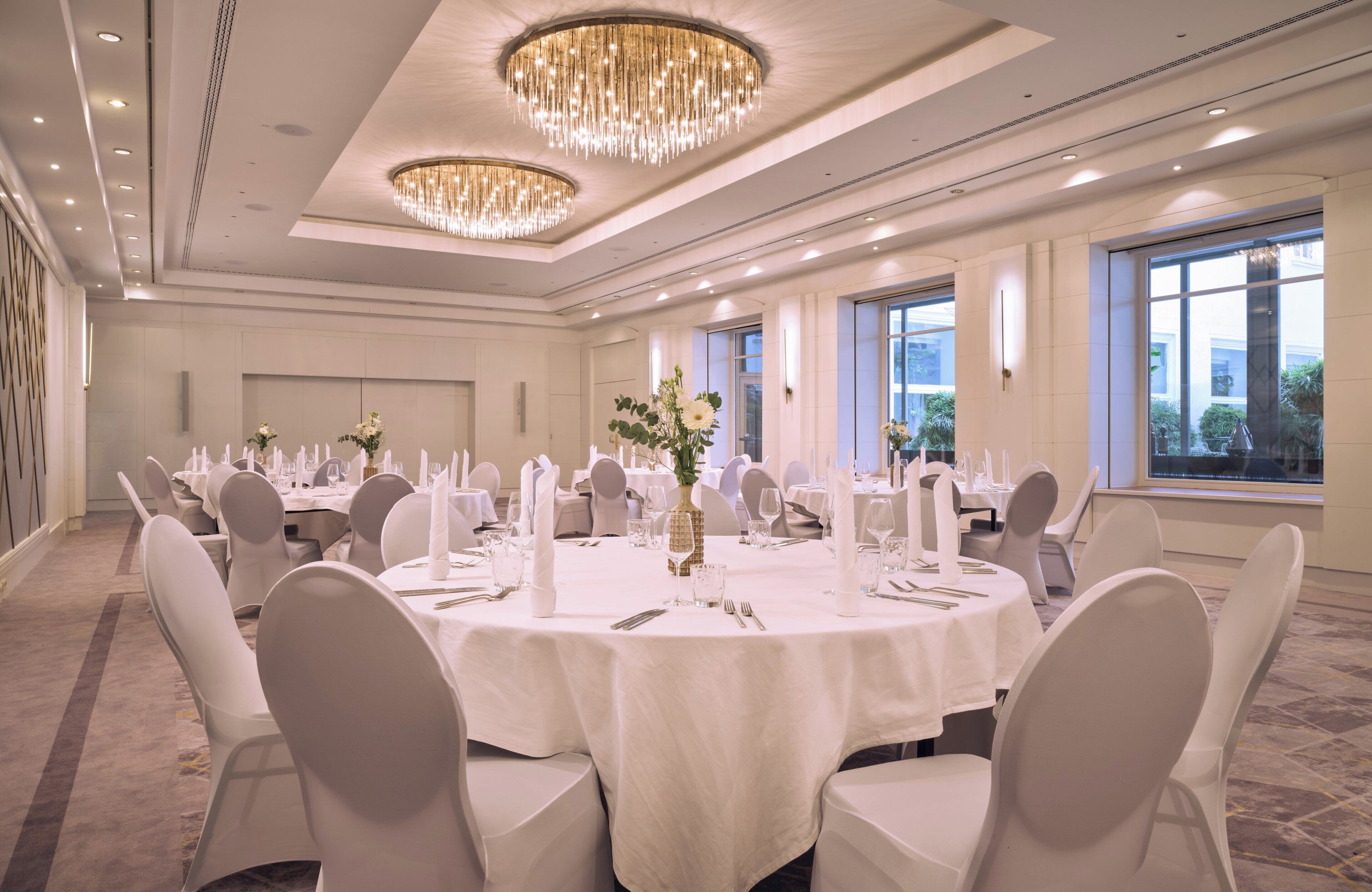 A banquet hall with round tables, white chairs, chandeliers, and large windows.