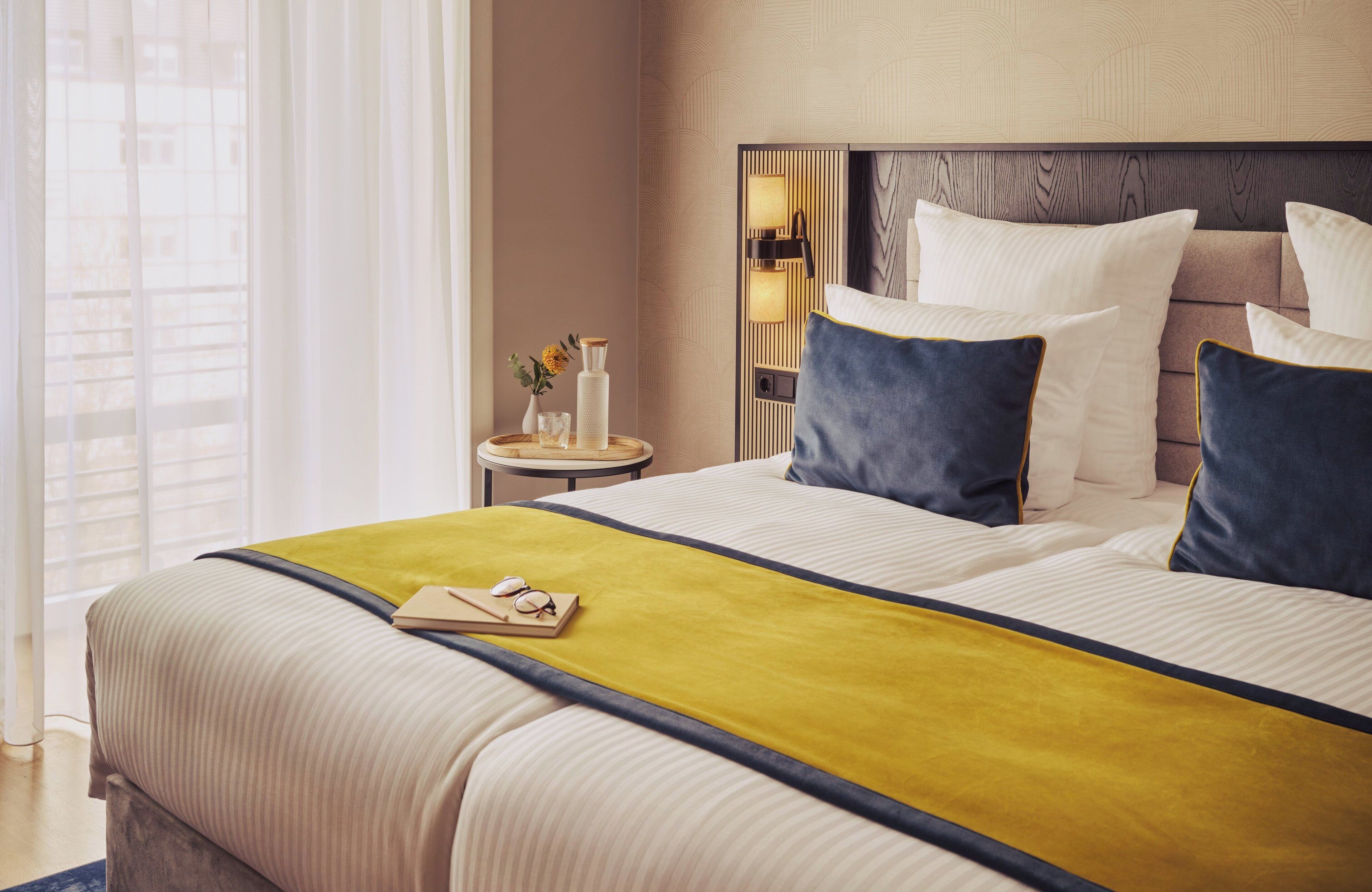 A hotel room with a large bed, a bedside table with a lamp, and a view of the city through the window.