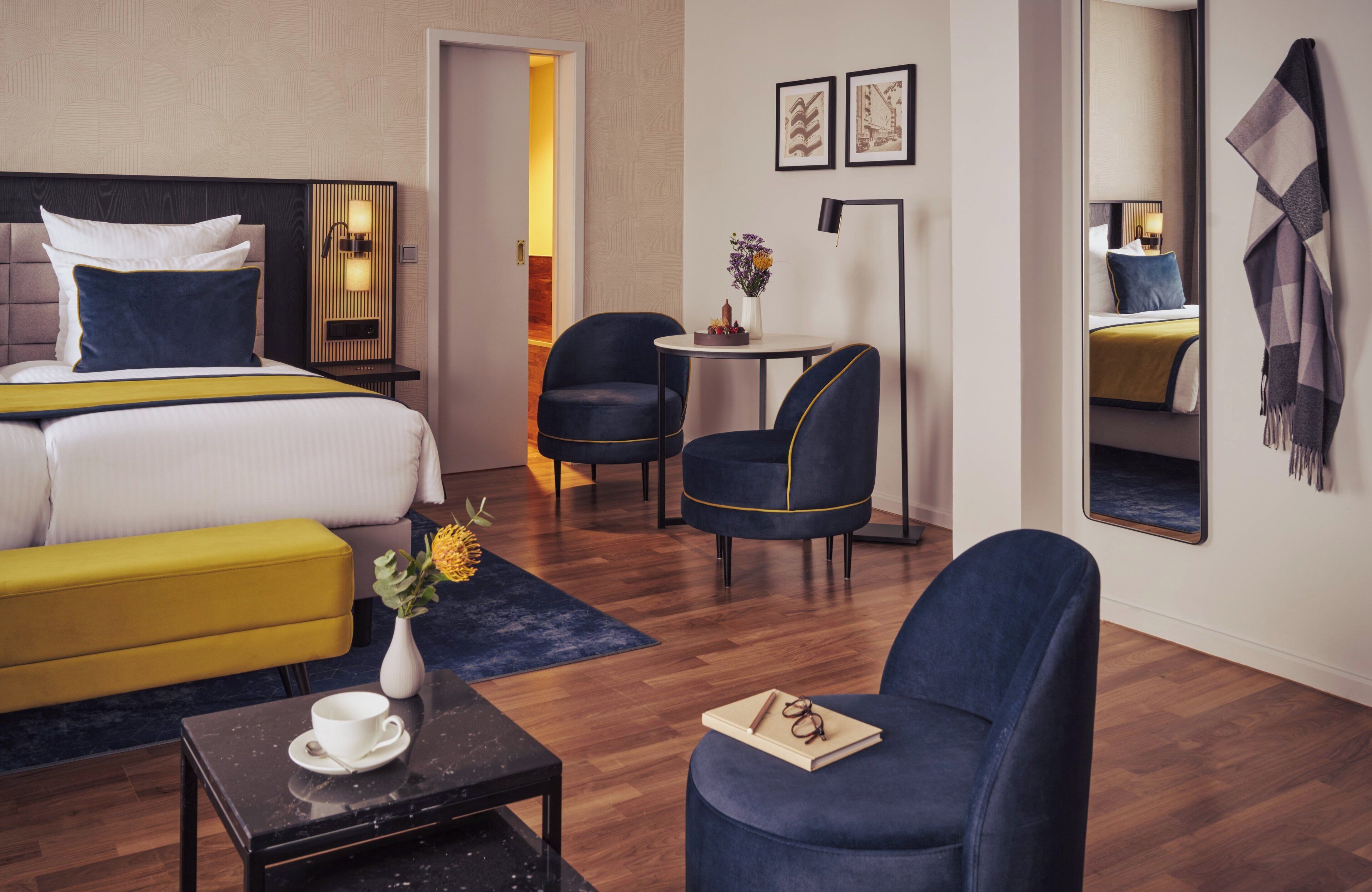 Une chambre d’hôtel avec un lit, un canapé jaune, une petite table avec une tasse et un livre, et deux fauteuils bleus.