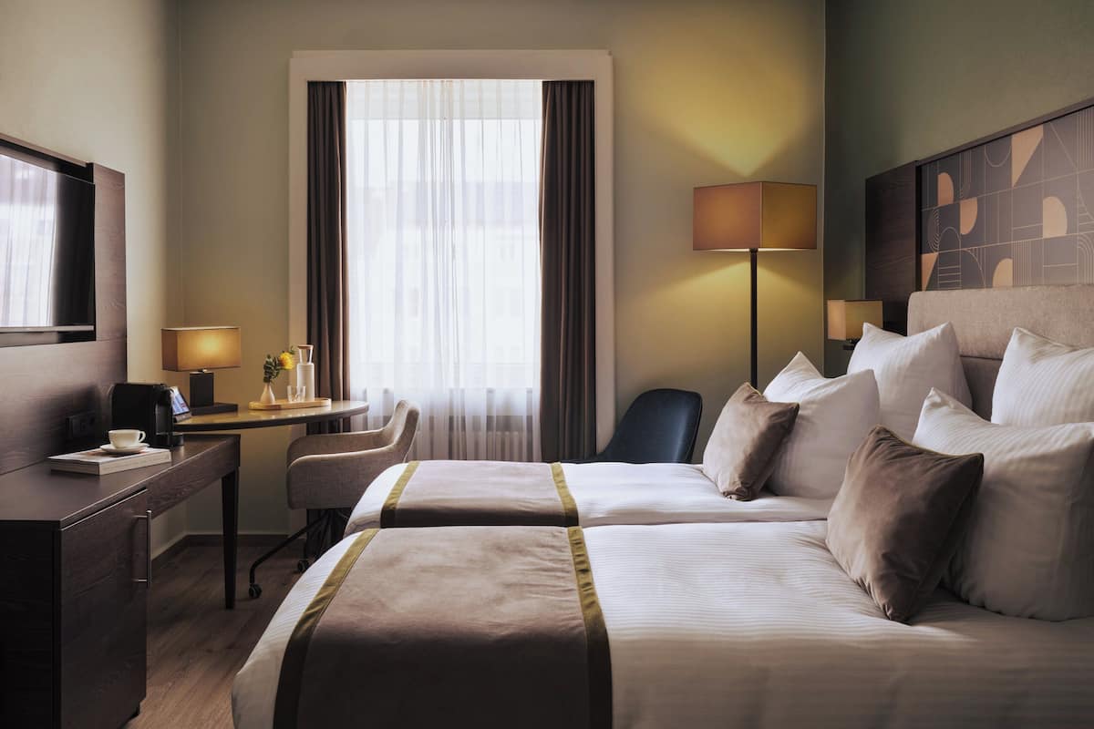 Quarto de hotel com duas camas, uma escrivaninha com abajur, uma cadeira e uma janela grande com cortinas.