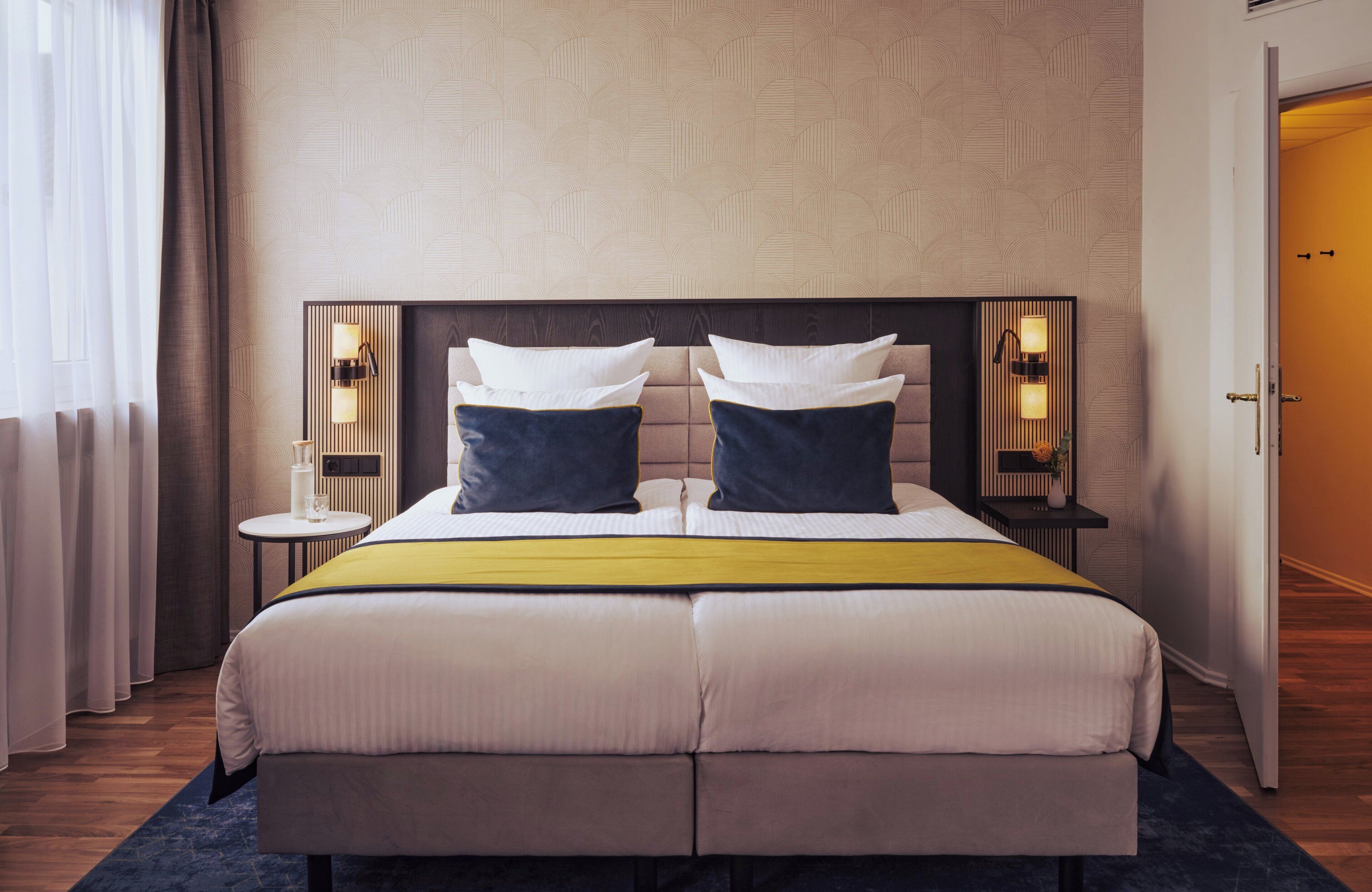 A double bed with a headboard, two bedside tables with lamps, a bedside table with a vase, and a door to the right.