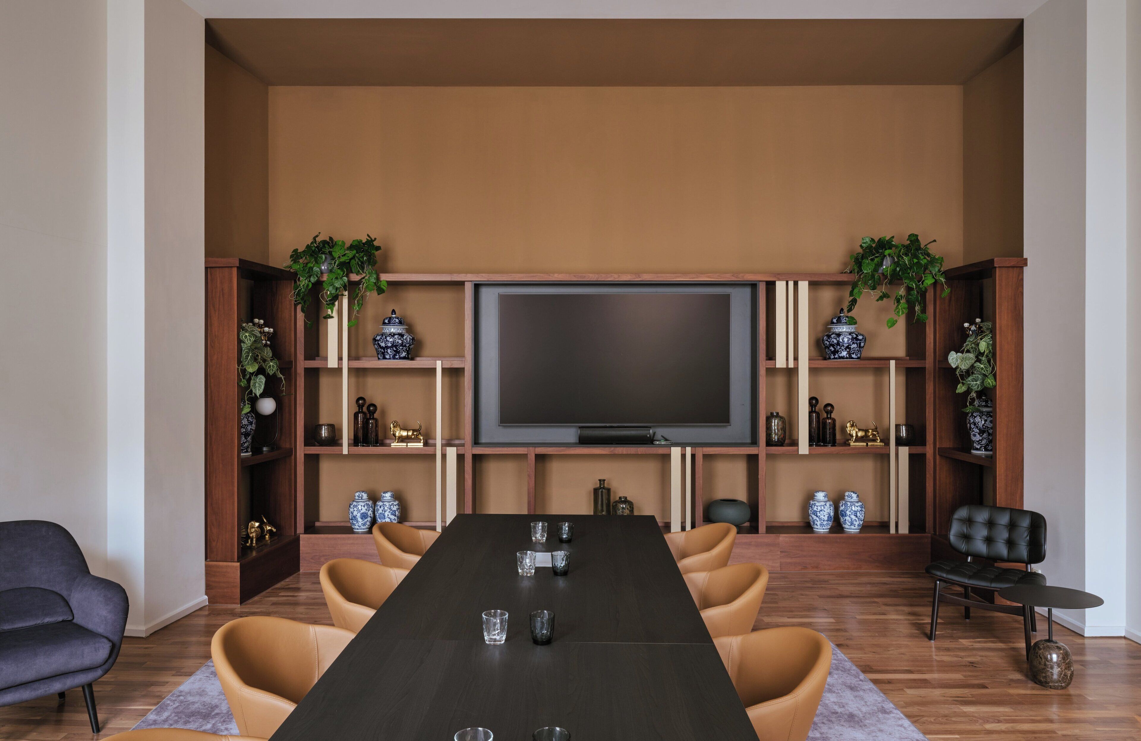 A modern living room with a long dining table, comfortable seating, a flat-screen TV, and decorative shelves with plants and vases.