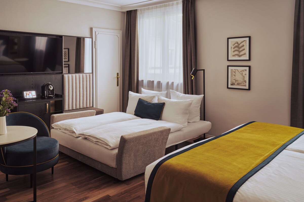 A hotel room with a bed, bedside table, chair, TV, and artwork on the wall.