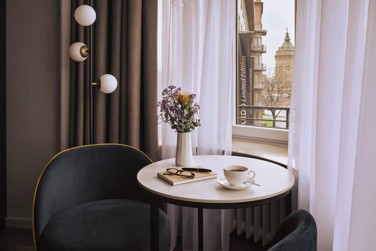 Quarto de hotel com uma mesa redonda, uma cadeira, um vaso com flores, uma chávena de café e um livro.