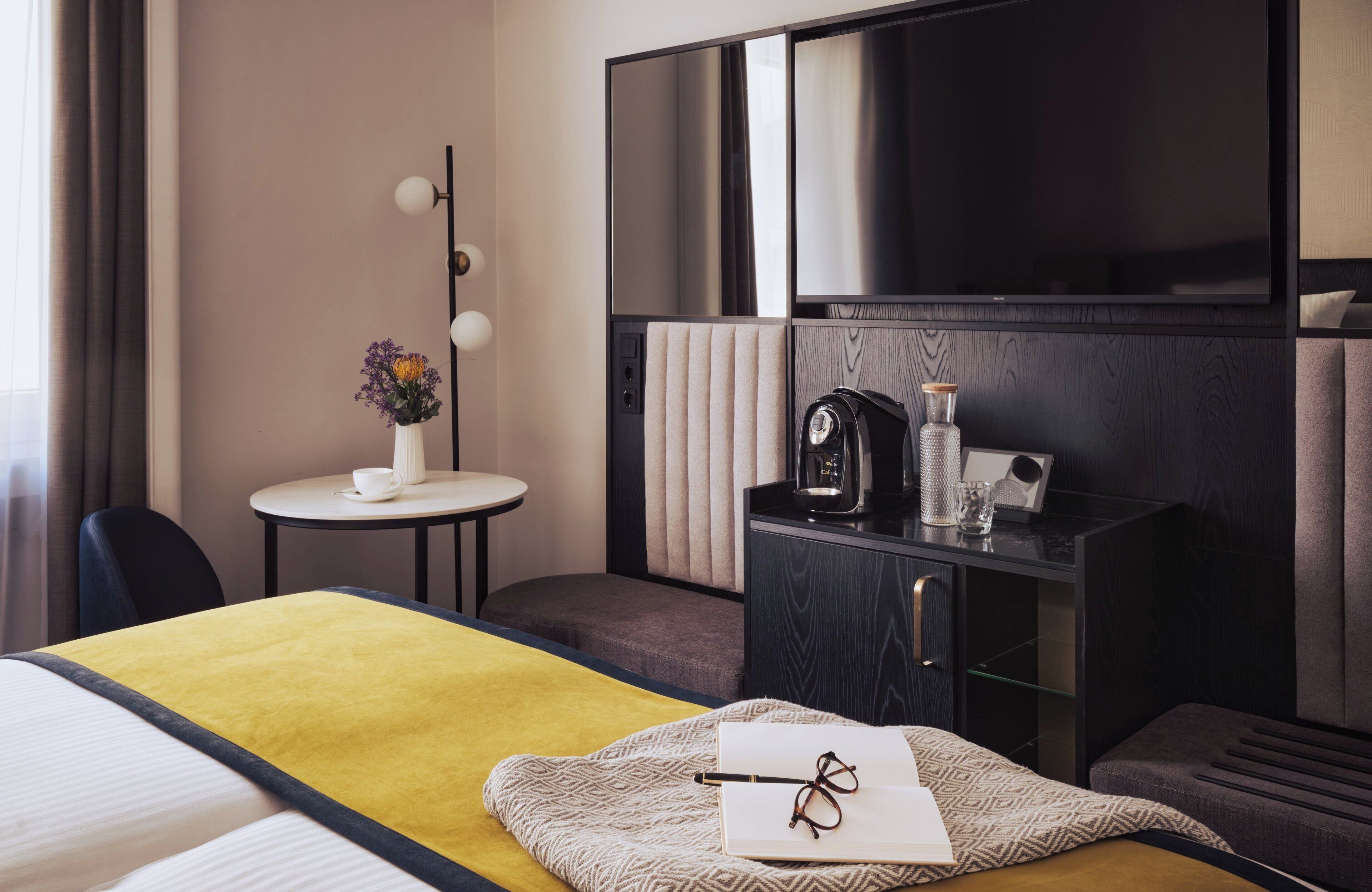 A hotel room with a bed, a bedside table with a lamp, a small table with a vase of flowers, a coffee maker, and a TV mounted on the wall.