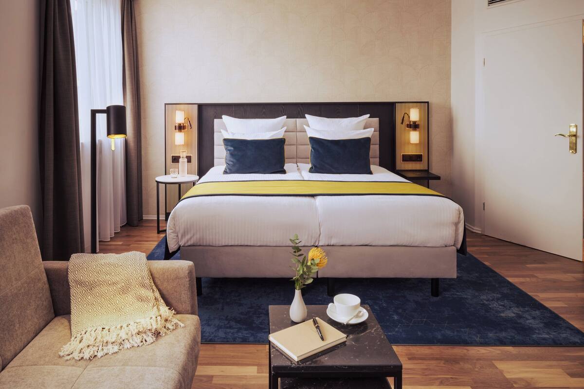 Ein Hotelzimmer mit einem großen Bett, zwei Nachttischen mit Lampen, einem Sofa, einem Couchtisch mit Buch und einer Blumenvase.