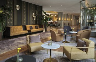 A modern lobby with a sofa, armchairs, and a coffee table. There are plants and a dining area in the background.