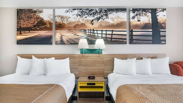 Una habitación de hotel con dos camas, una mesita de noche con dos lámparas y un cuadro grande con un paisaje sobre las camas.
