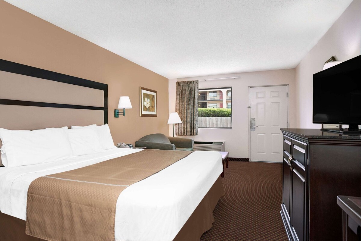 A hotel room with a bed, bedside table, lamp, TV, and a window with a view of buildings.