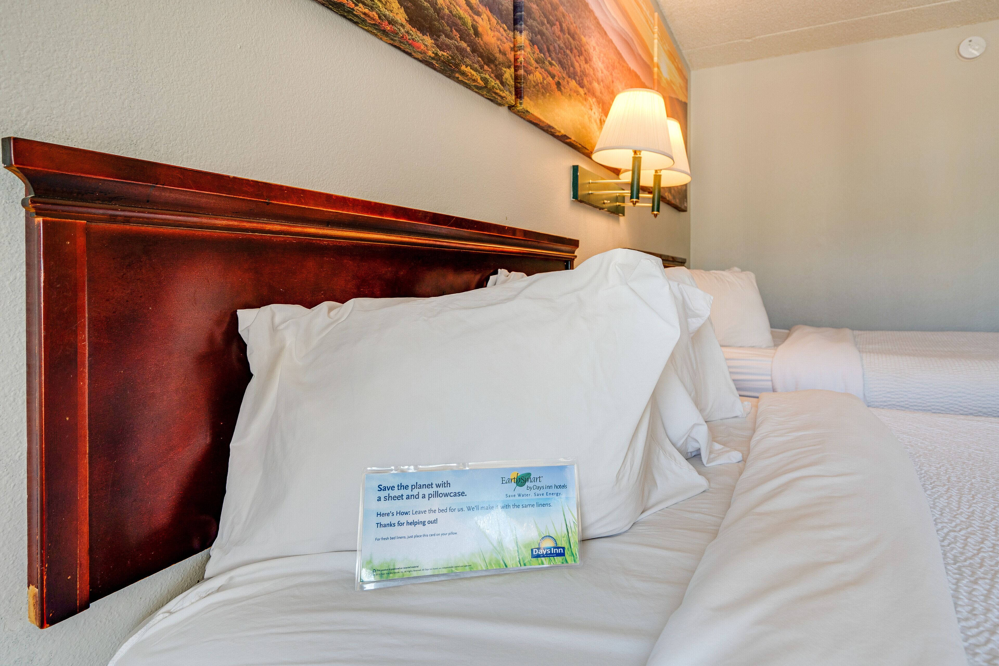 A hotel room with a wooden headboard, a bed with white linens, a wall-mounted lamp, and a sign promoting eco-friendly practices.