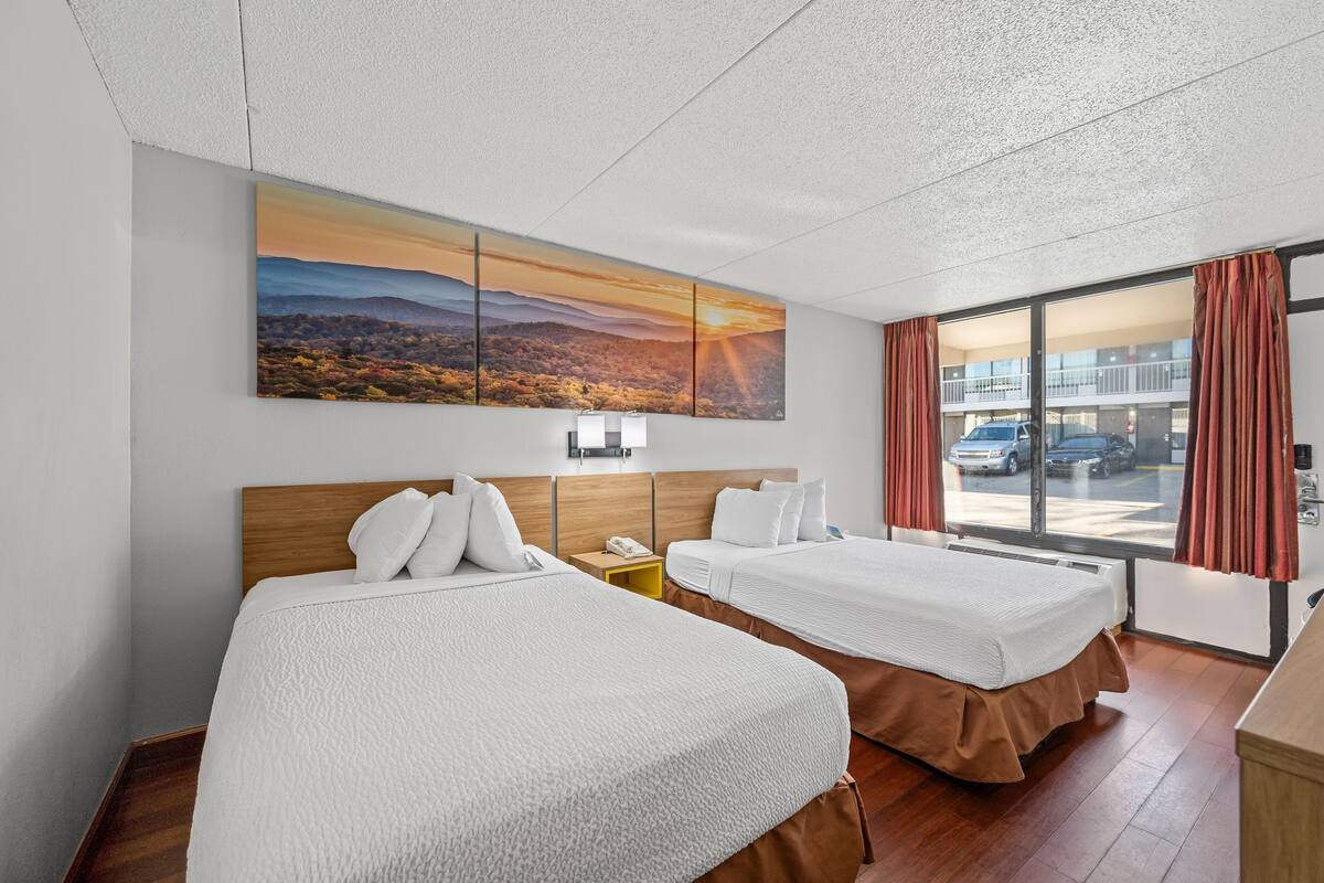 A hotel room with two beds, a large window with curtains, a painting of a sunset, and a view of the street outside.