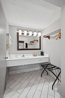 A modern bathroom with a white sink, a large mirror with lights, a towel rack, and a black chair.
