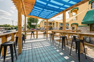 An outdoor deck with wooden tables and black bar stools, overlooking a street with parked cars and a building with a yellow facade.