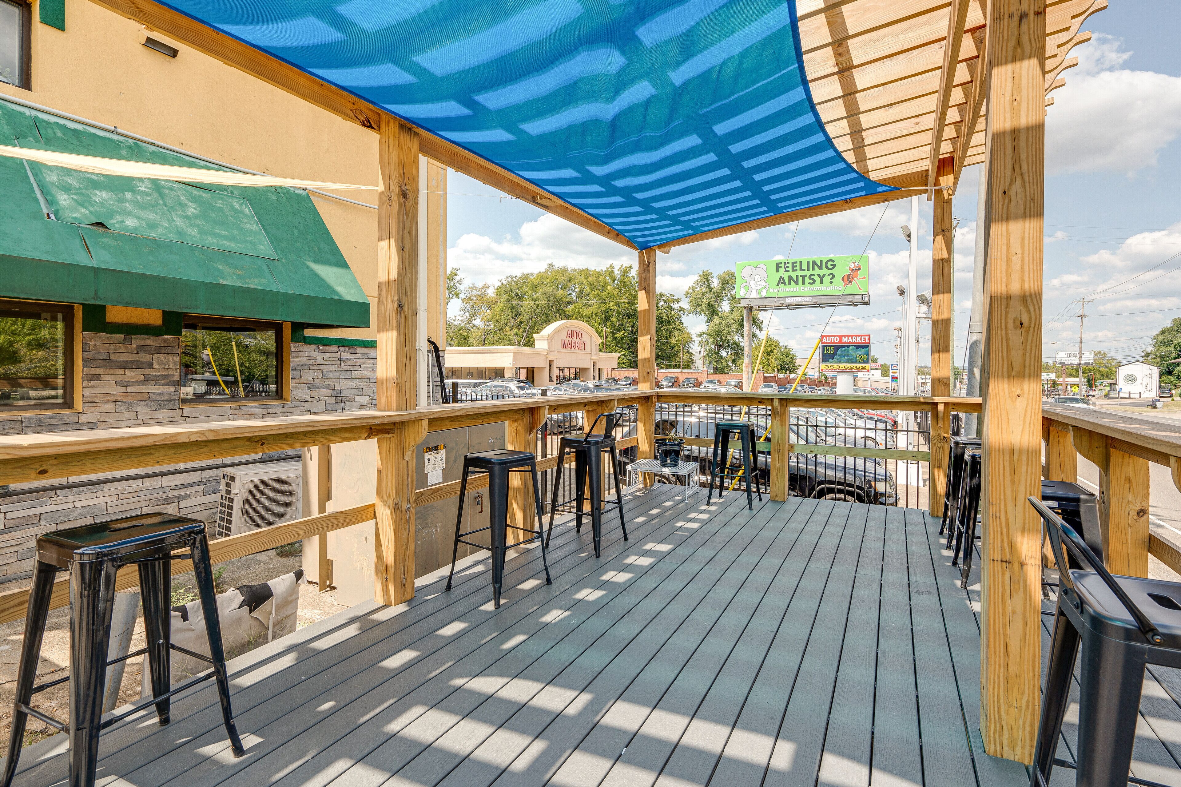 An outdoor deck with wooden railings and black bar stools, overlooking a parking lot and a building with a green awning.