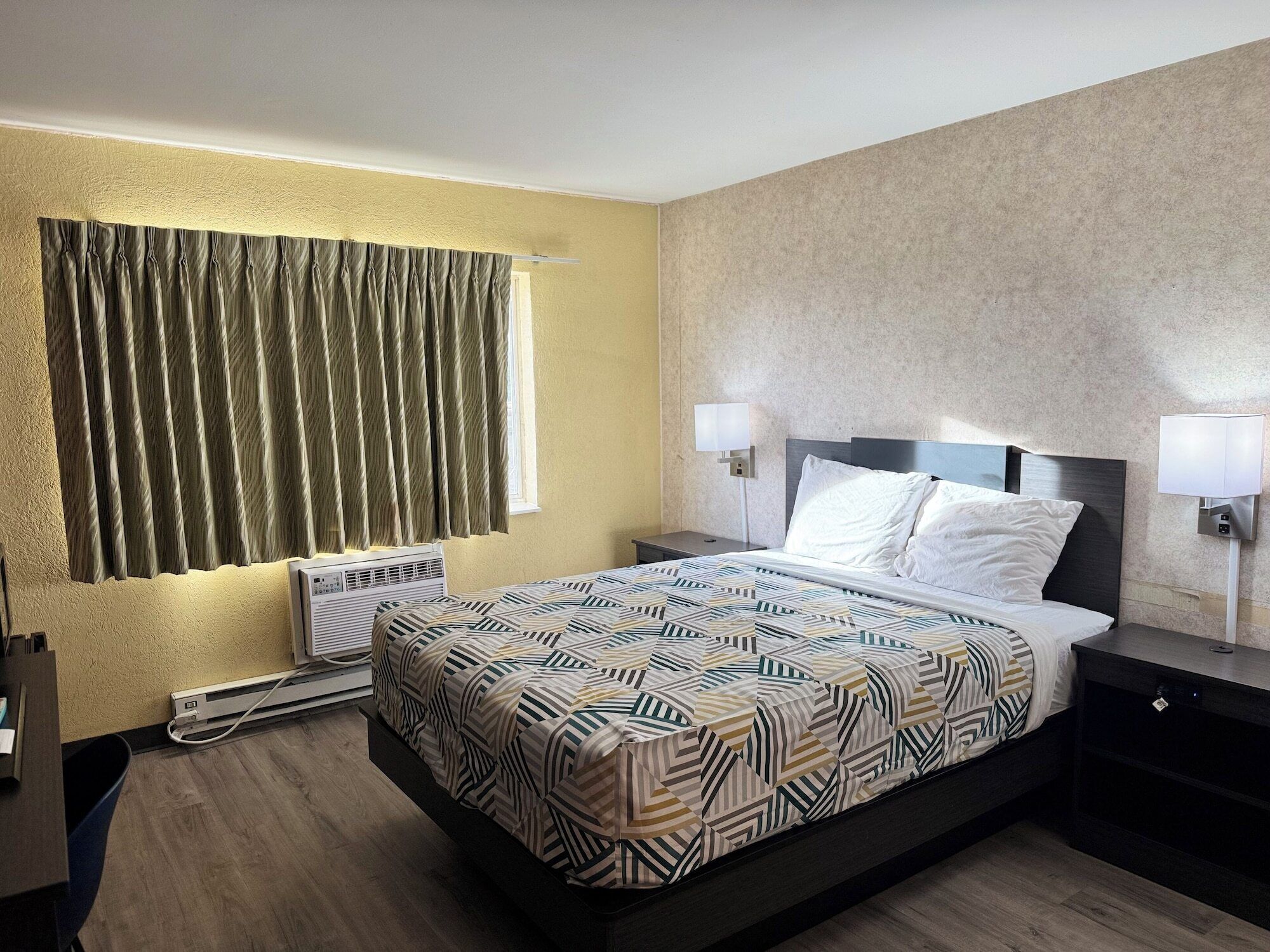 A hotel room with a bed, two bedside lamps, a window with curtains, an air conditioning unit, and a desk with a chair.