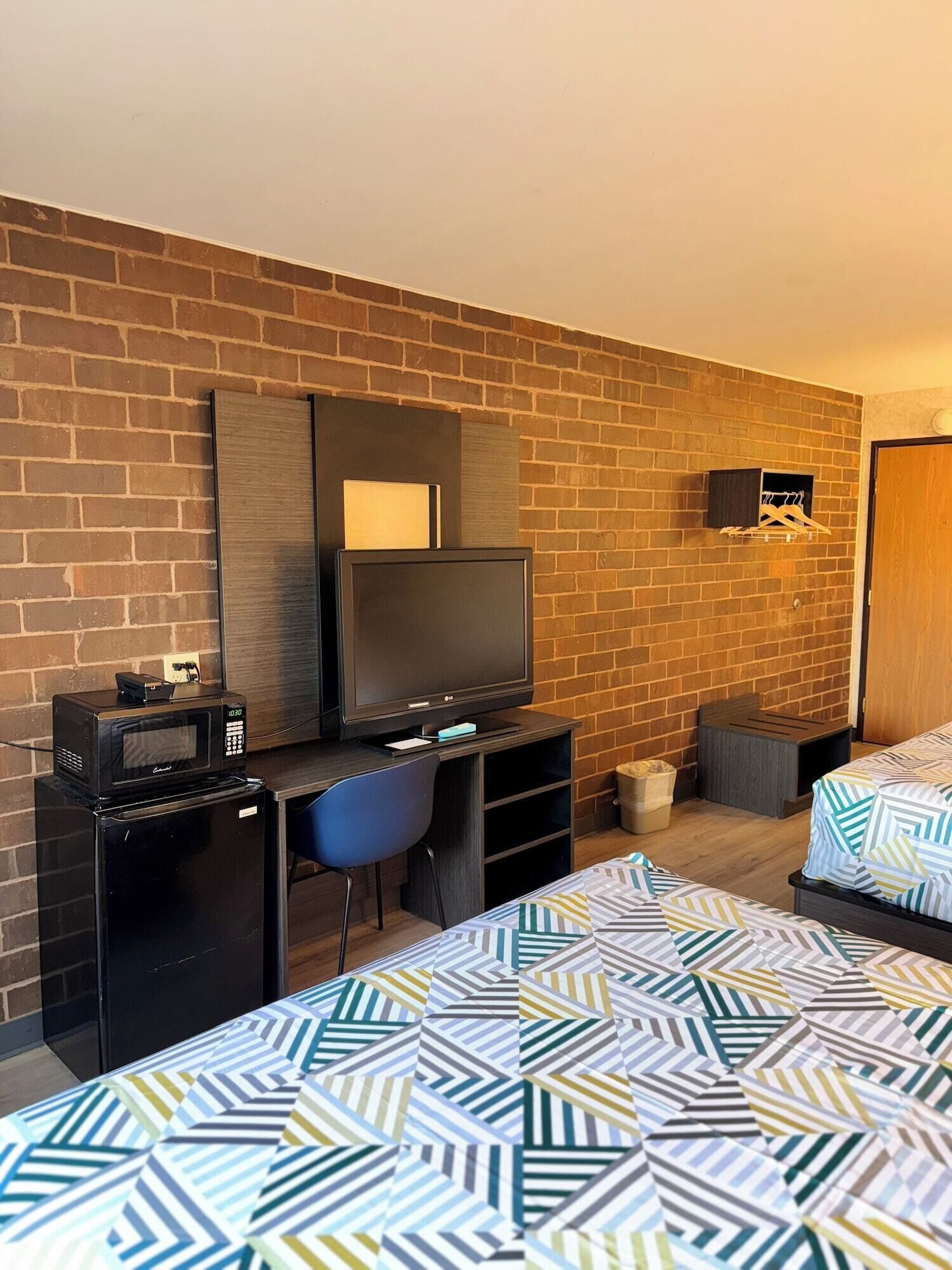 Habitación de hotel con pared de ladrillo, un televisor de pantalla plana, microondas, escritorio con silla y dos camas con ropa de cama estampada.
